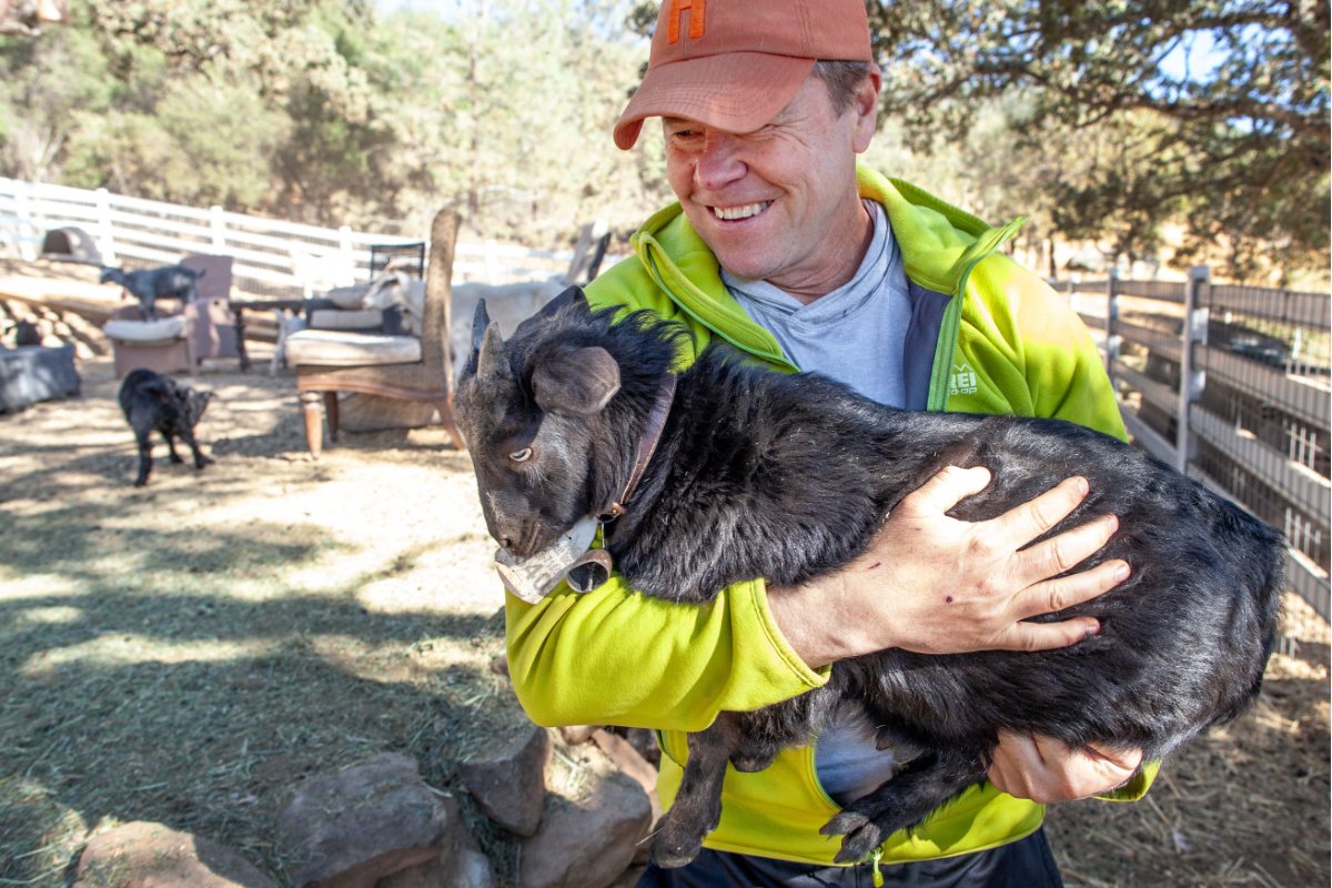 A farmer wearing an orange gap smiles as he holds a small black goat
